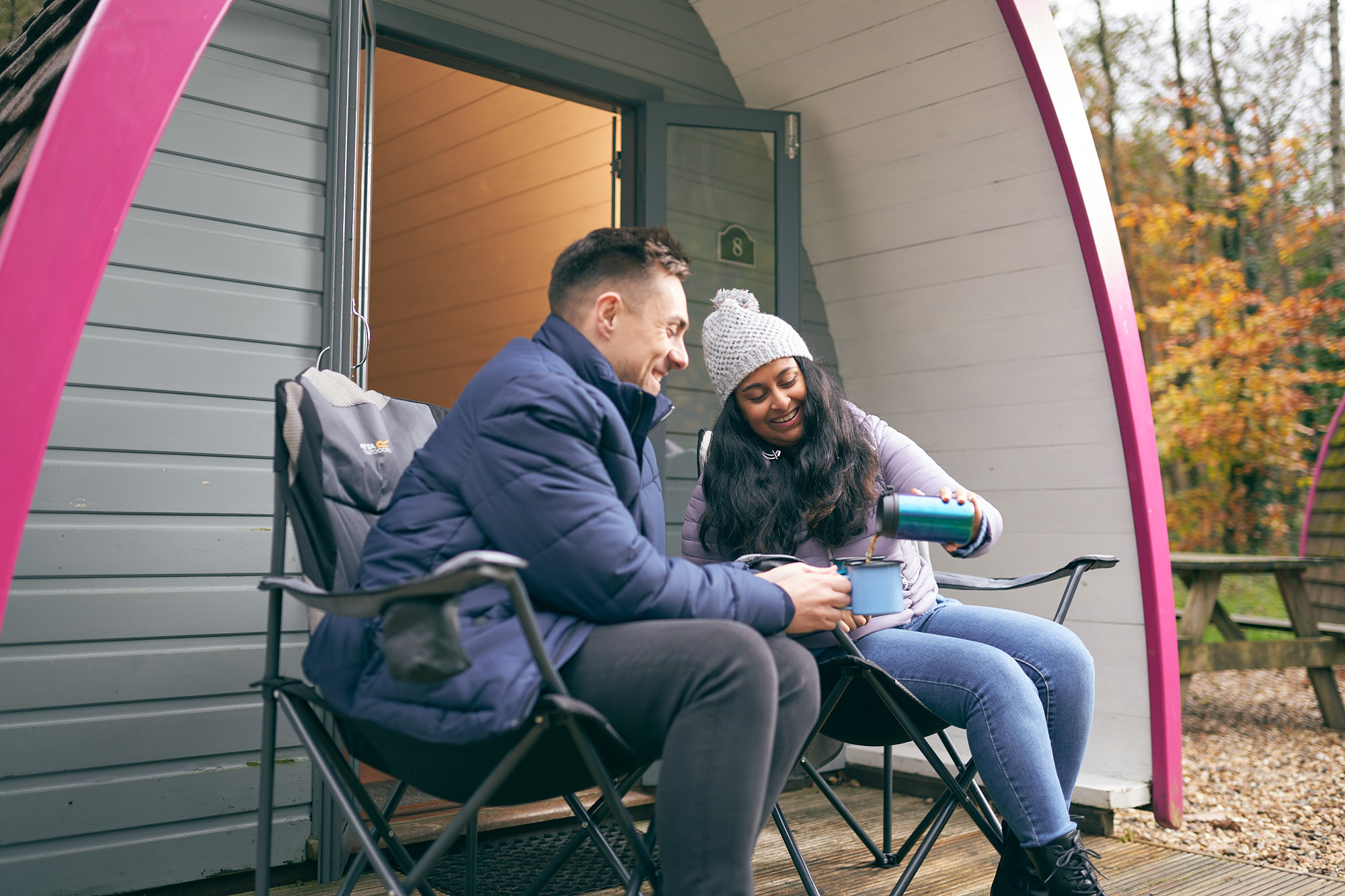 Dyn a dynes yn eistedd tu allan pod glampio yn gwenu. Mae'r ddynes yn arllwys diod dwym i fwg iddynt fwynhau.