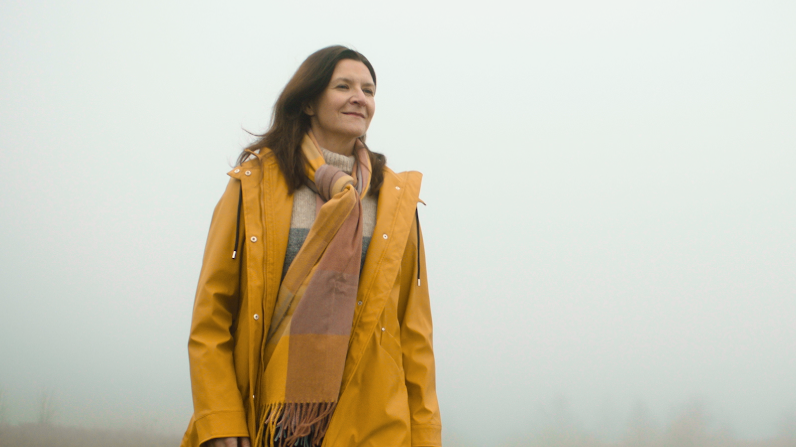 Dynes mewn cot a sgarff melyn yn gwenu ac edrych i'r pellter yn yr awyr agored