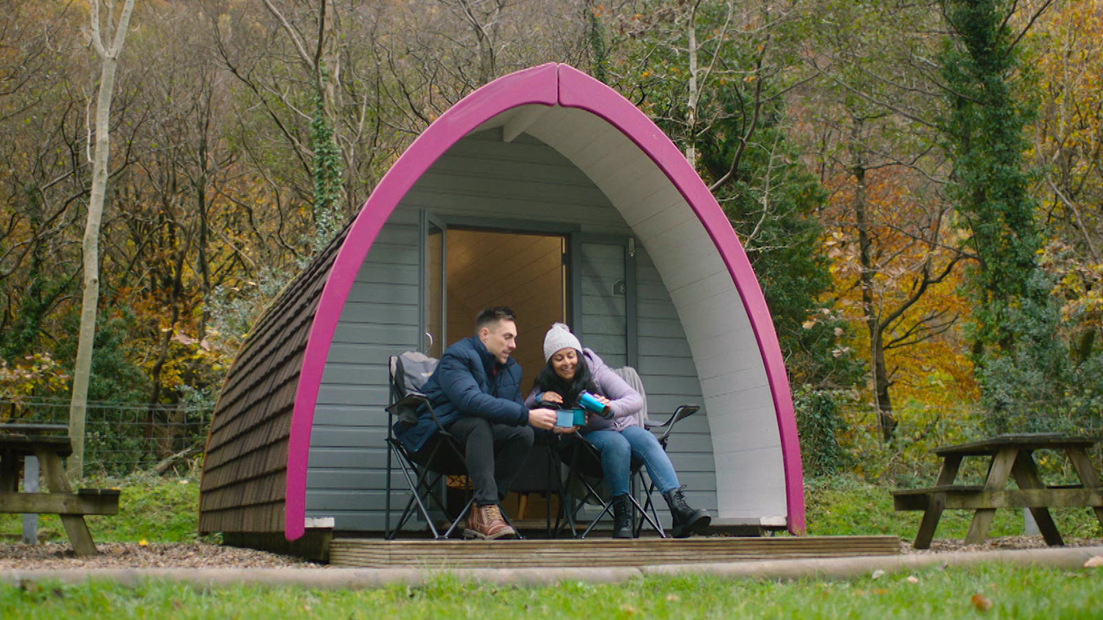 Dyn a dynes yn eistedd tu allan pod glampio yn y goedwig yn mwynhau diod twym.